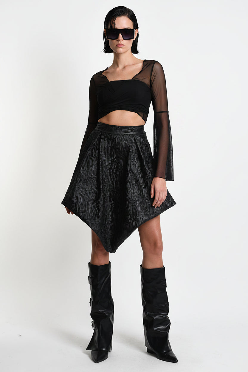 A model wearing a black gothic-style outfit consisting of a sheer long-sleeve crop top, a textured black pleated mini skirt, and black knee-high boots, photographed against a white studio background.