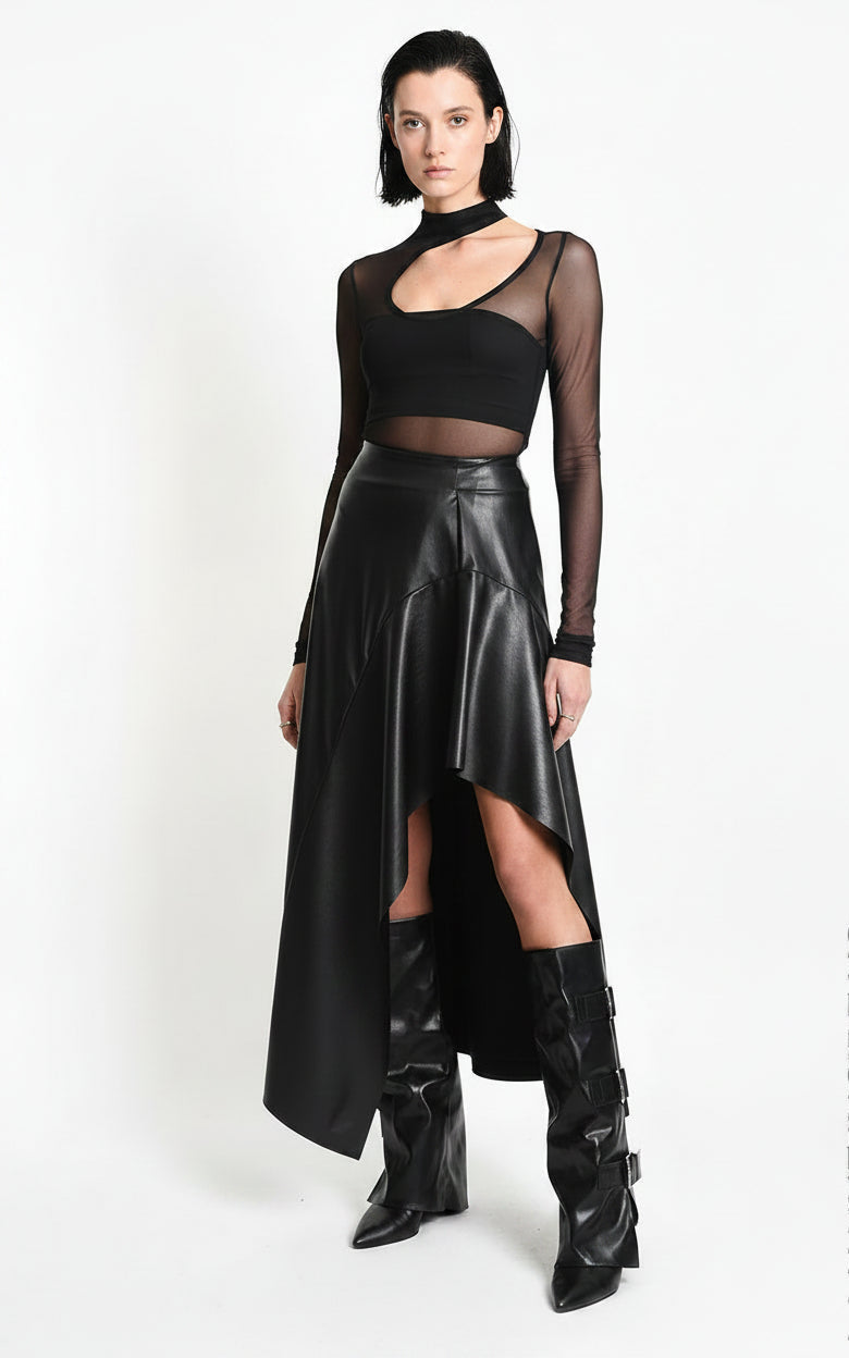 A model wearing a black asymmetrical high-low faux leather skirt with a sheer long-sleeve crop top and black leather boots, standing against a white studio background.