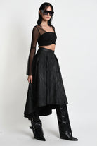 A model wearing a black fashion ensemble consisting of a sheer long-sleeve crop top, a textured black high-waisted skirt, and black leather boots, photographed against a white studio background.
