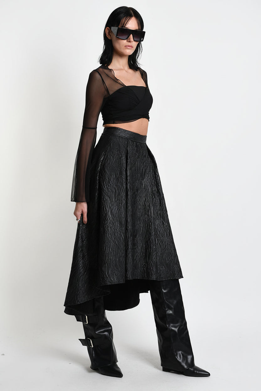 A model wearing a black fashion ensemble consisting of a sheer long-sleeve crop top, a textured black high-waisted skirt, and black leather boots, photographed against a white studio background.