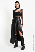 A model wearing a black asymmetrical high-low faux leather skirt with a sheer long-sleeve crop top and black leather boots, standing against a white studio background.